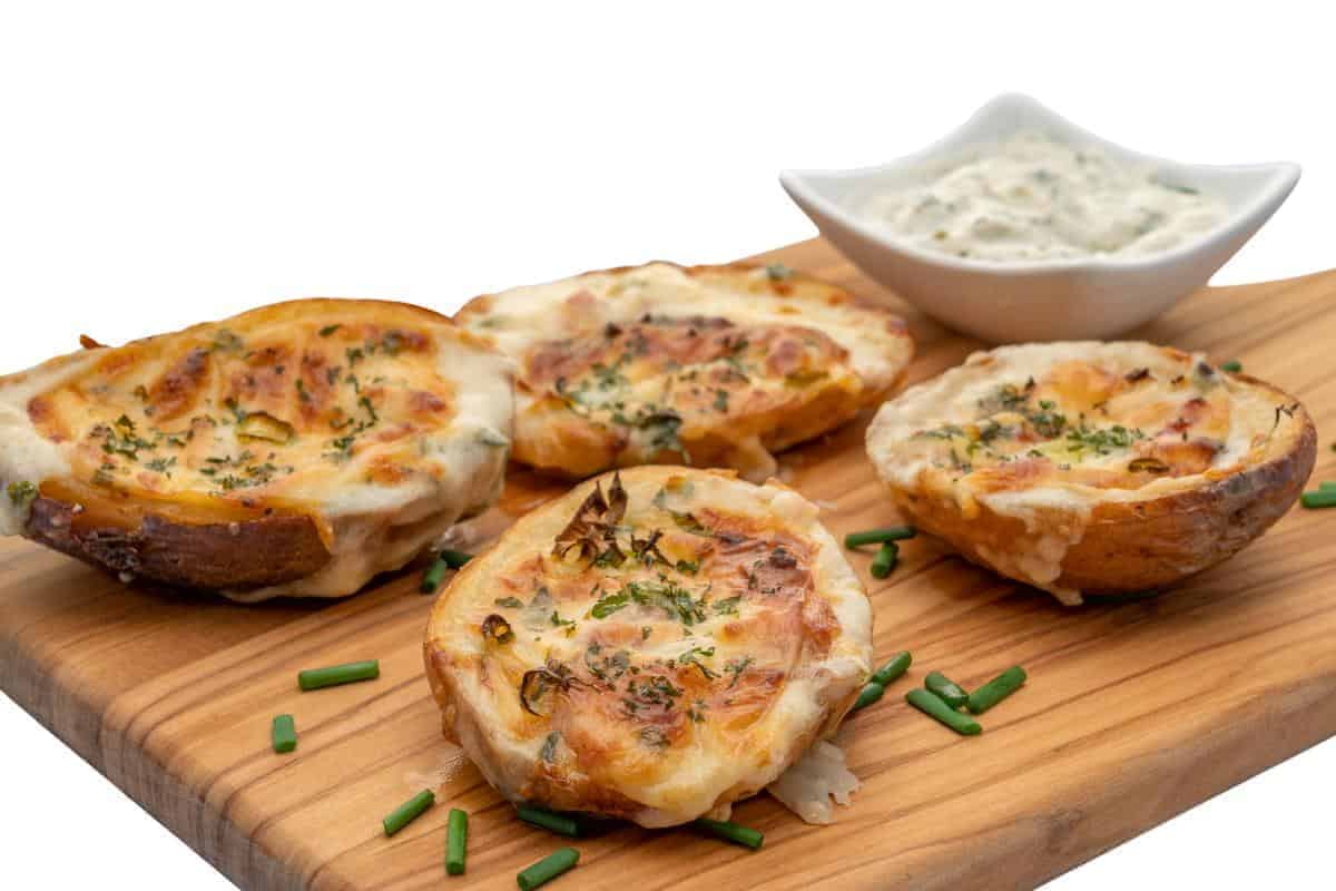 Four loaded potato skins topped with melted cheese and herbs are arranged on a wooden board, garnished with chopped chives. A small bowl of creamy dipping sauce sits nearby. This easy loaded potato skins recipe is perfect for families.