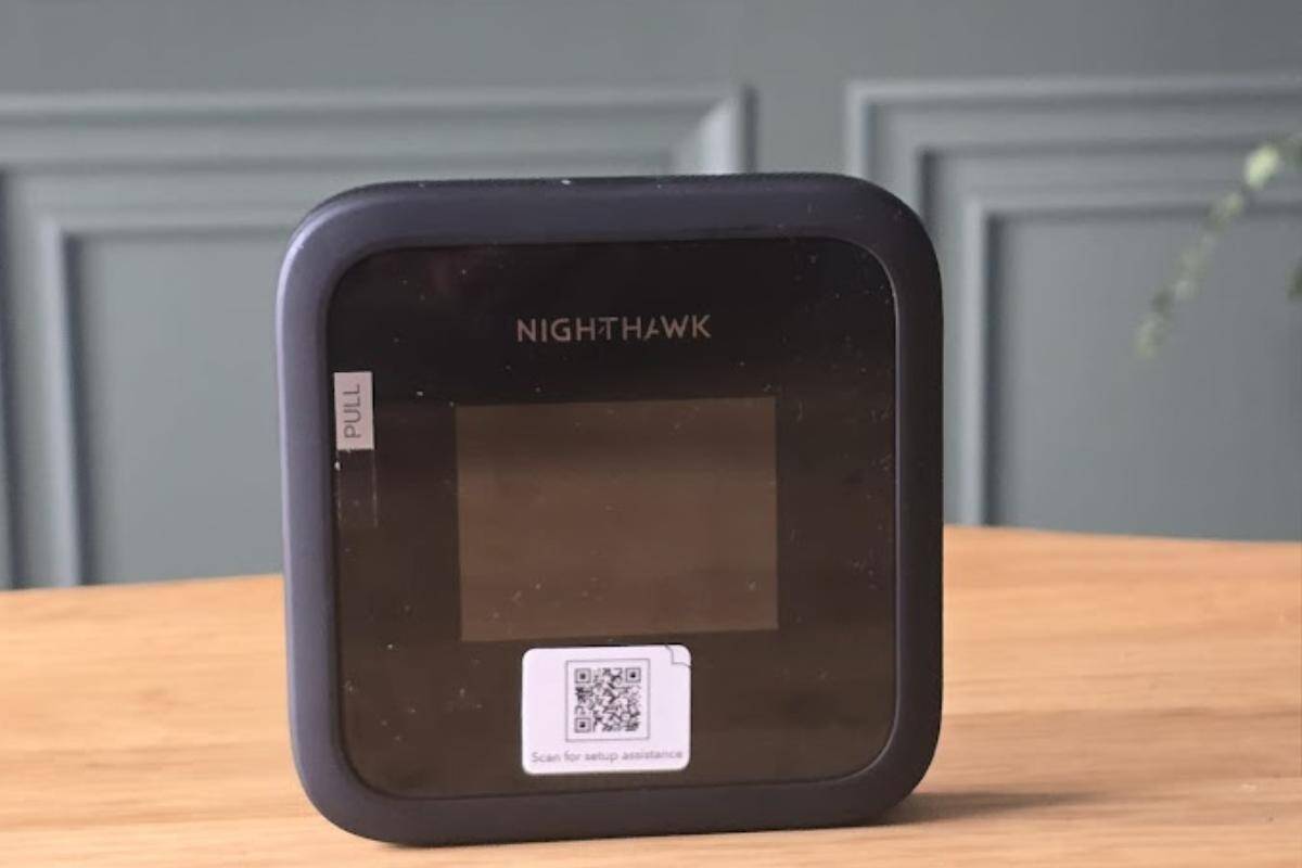 A small, black rectangular mobile WiFi hotspot labeled "NIGHTHAWK" sits on a wooden surface. With a screen in the center and setup QR code below, it's perfect for travelers wanting to stay connected in Europe. The background features a gray wall with molding detail.