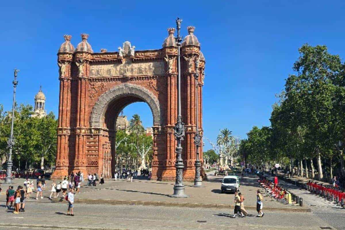 The Arc de Triomf, a large red-brick triumphal arch in Barcelona, stands under a clear blue sky, surrounded by trees and people walking and cycling—an ideal spot to pause during your Barcelona city tour bus adventure.