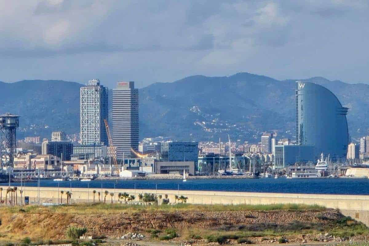 Skyline of Barcelona featuring tall modern buildings, including the sail-shaped W Hotel by the waterfront—where many travelers from Alcudia to Barcelona arrive via Balearia Ferry—with mountains in the background and a partly cloudy sky above.
