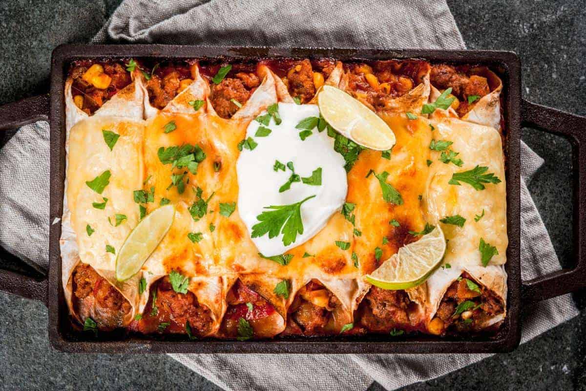 A casserole dish filled with Easy Beef Enchiladas topped with melted cheese, sliced lime wedges, a dollop of sour cream, and fresh cilantro. The dish sits on a gray napkin on a dark countertop.