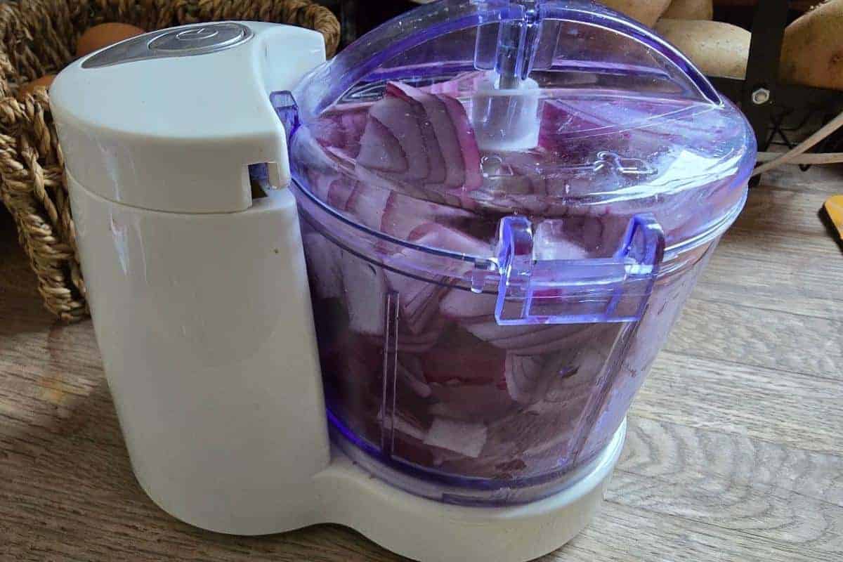 A food processor on a wooden surface is filled with chopped red onions, perfect for starting your chicken bhuna recipe. A basket with eggs and potatoes is visible in the background.