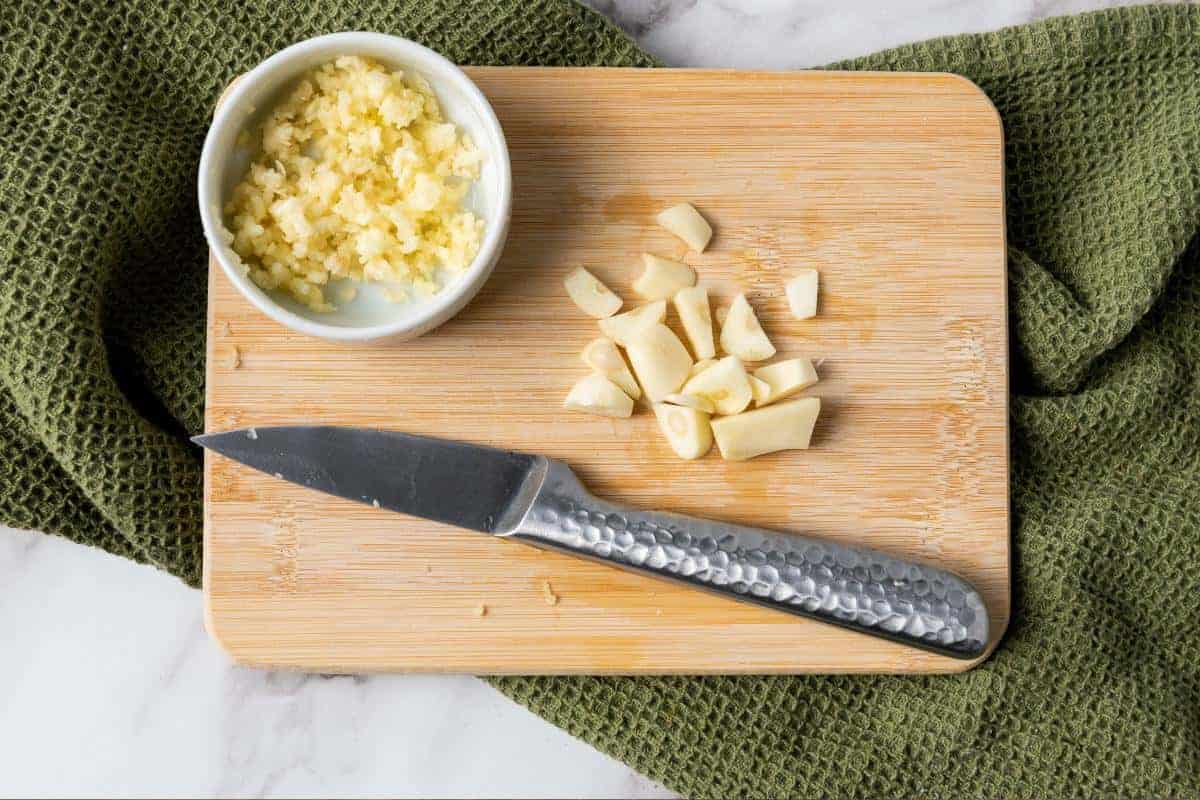A cutting board with chopped garlic, a paring knife, a small bowl of minced garlic, and a green textured cloth underneath on a marble surface—perfect prep for delicious rice or an easy side dish like Garlic Rice.