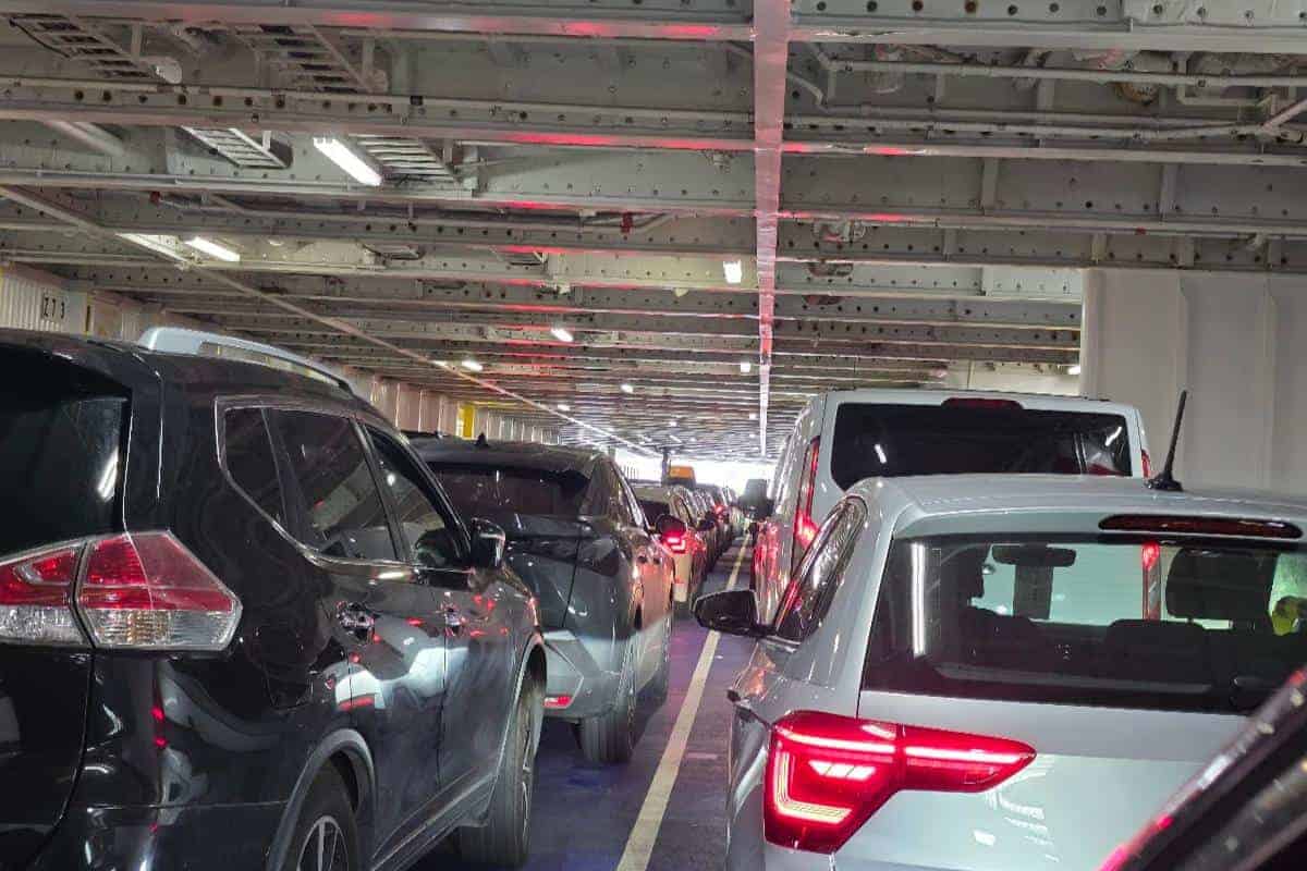 Rows of cars are parked closely together inside the lower deck of the Dover to Calais Ferry, with overhead metal beams and fluorescent lights on the ceiling. Red brake lights illuminate the scene, indicating vehicles are stationary but this could be scary and cause sensory overload on the ferry with autism.