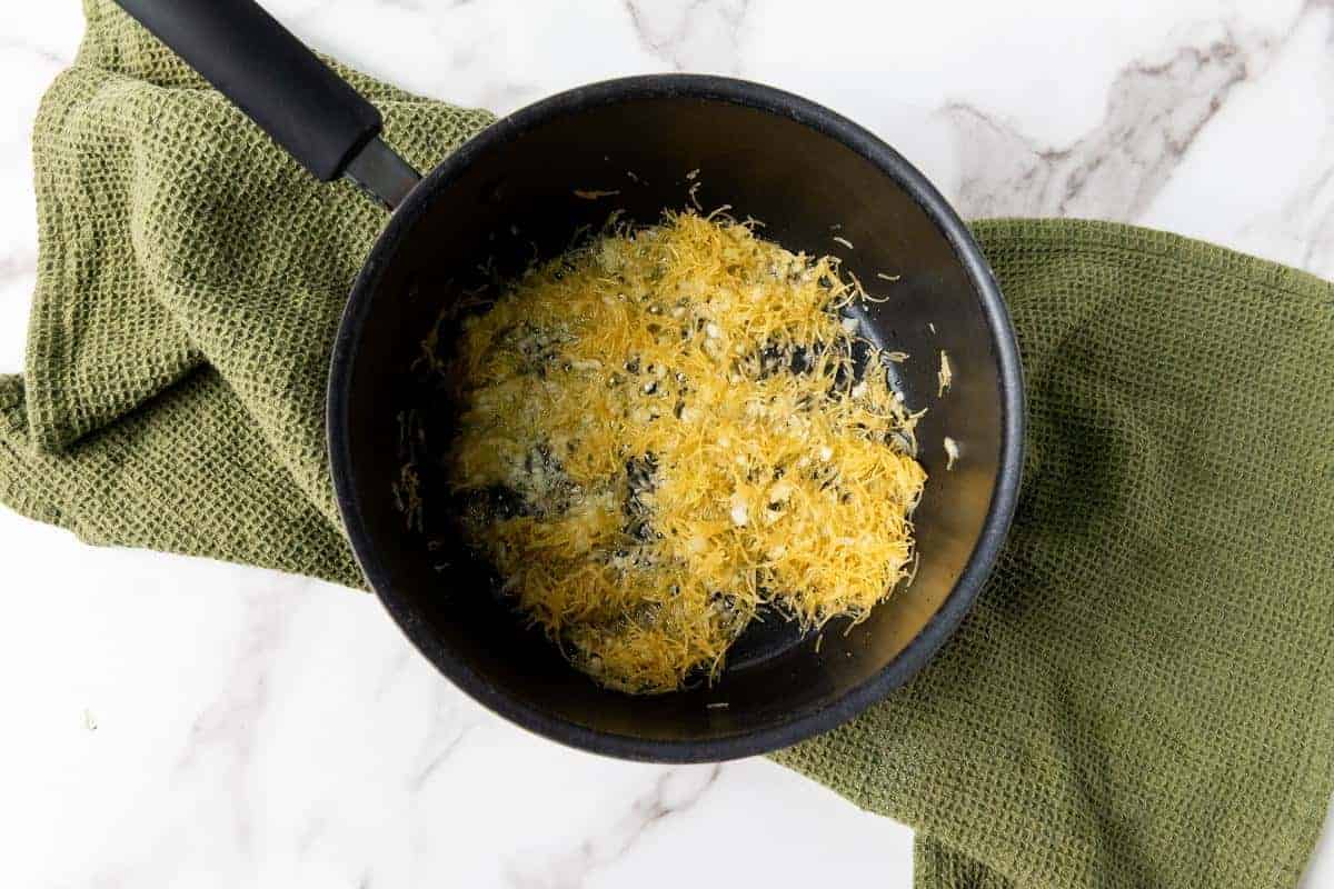 A black saucepan containing shredded golden-brown food, perfect for an easy garlic rice side dish, sits on a green textured cloth atop a white marble surface.