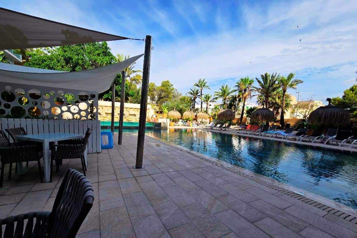 Outdoor swimming pool lined with lounge chairs and straw umbrellas at Bahia del Sol, Santa Ponsa, surrounded by palm trees under a blue sky; shaded seating area with tables and chairs in the foreground, perfect for an all-inclusive getaway.