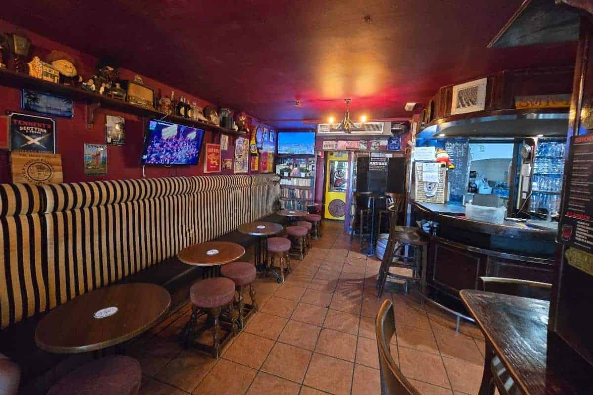 A cozy all-inclusive pub with striped cushioned seating, round wooden tables, and stools. The bar area is on the right, and the walls are decorated with posters, memorabilia, and shelves of books. The lighting is warm and inviting.