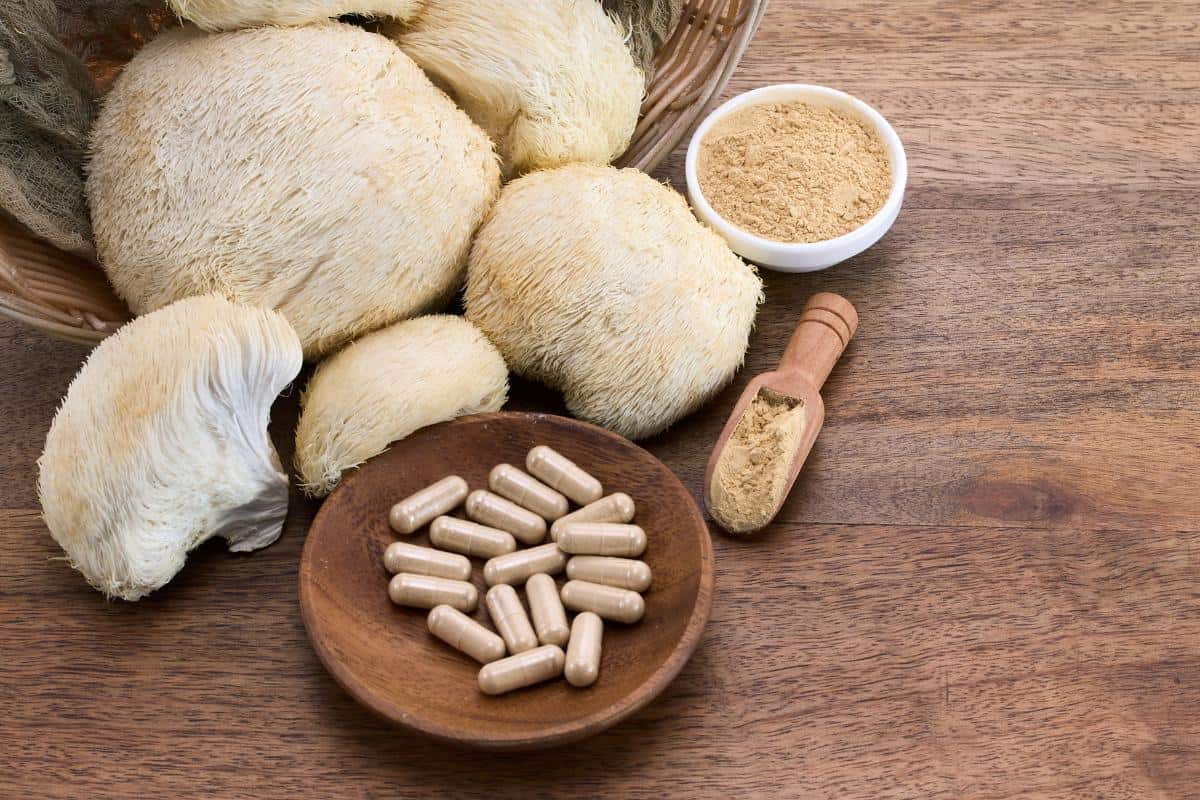 Lion’s Mane mushrooms on a wooden surface alongside a bowl of capsules, a small bowl of powdered lion's mane for weight loss supplement, and a wooden scoop with powder.