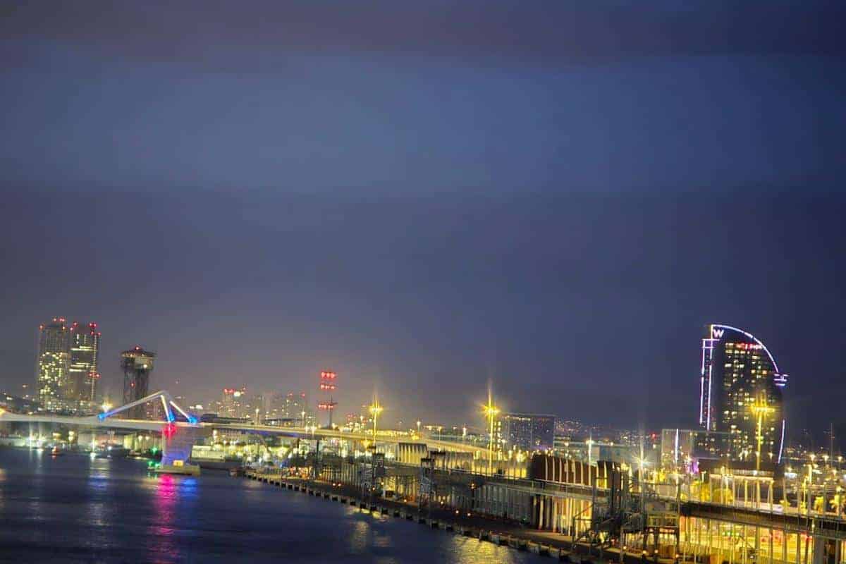 Nighttime cityscape featuring illuminated buildings and a waterfront with piers, evoking the allure of an overnight journey. Colorful lights reflect on the water, and a tall, curved building stands out on the right against the dark sky.