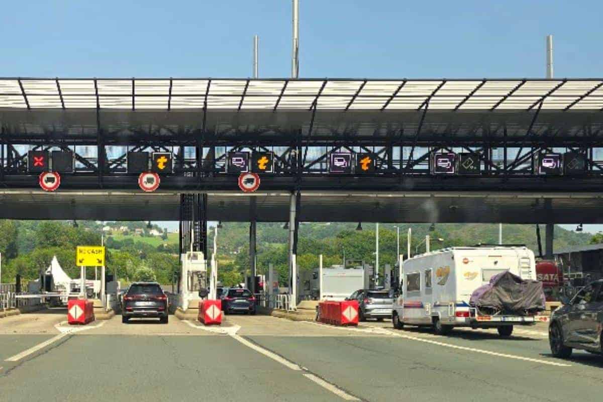 Cars and a camper van approach a French toll road booth, with illuminated electronic signs above each lane indicating payment types, including Emovis tag access. This is one of the toll roads in France.