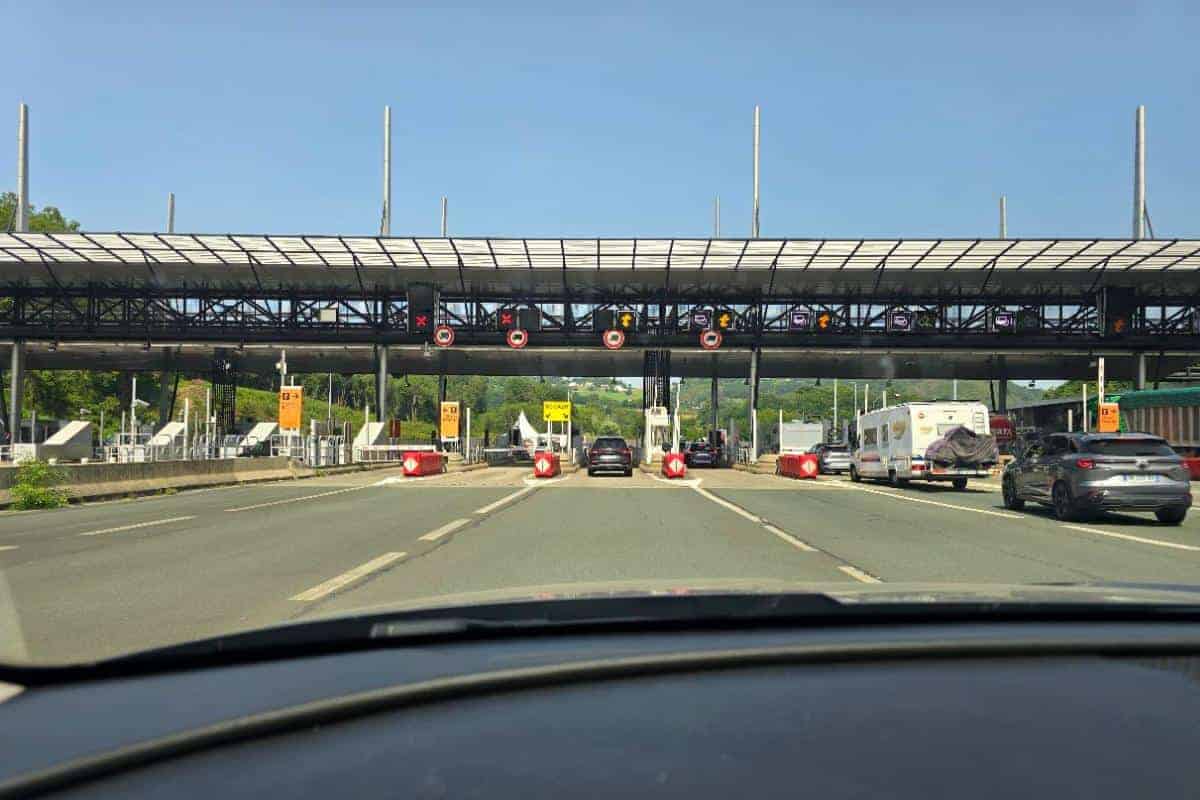 View from a car approaching a French toll booth, with several closed lanes marked by red lights and one open lane indicated by a green arrow. Cars line up to pass through—consider using an Emovis tag for quicker access on French toll roads.