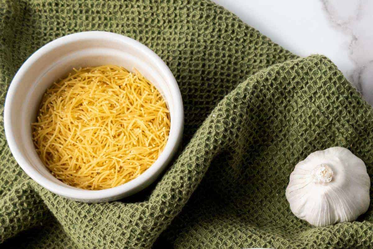 A white bowl filled with uncooked vermicelli noodles sits on a textured green cloth next to a whole garlic bulb on a white marble surface—perfect ingredients for an easy Garlic Rice side dish.