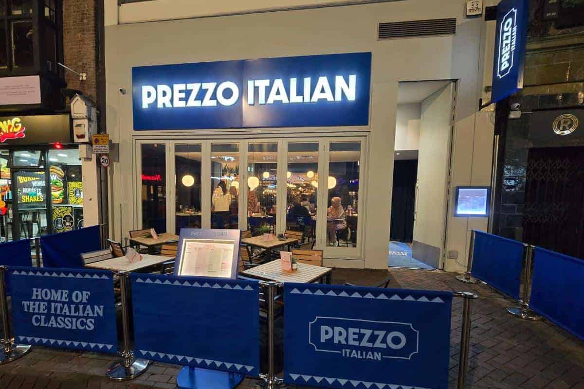 A brightly lit Prezzo Italian Nottingham restaurant at night, with outdoor seating and blue barriers displaying the restaurant’s name and “Home of the Italian Classics.” Diners are visible inside through large windows. Perfect for your next restaurant review.