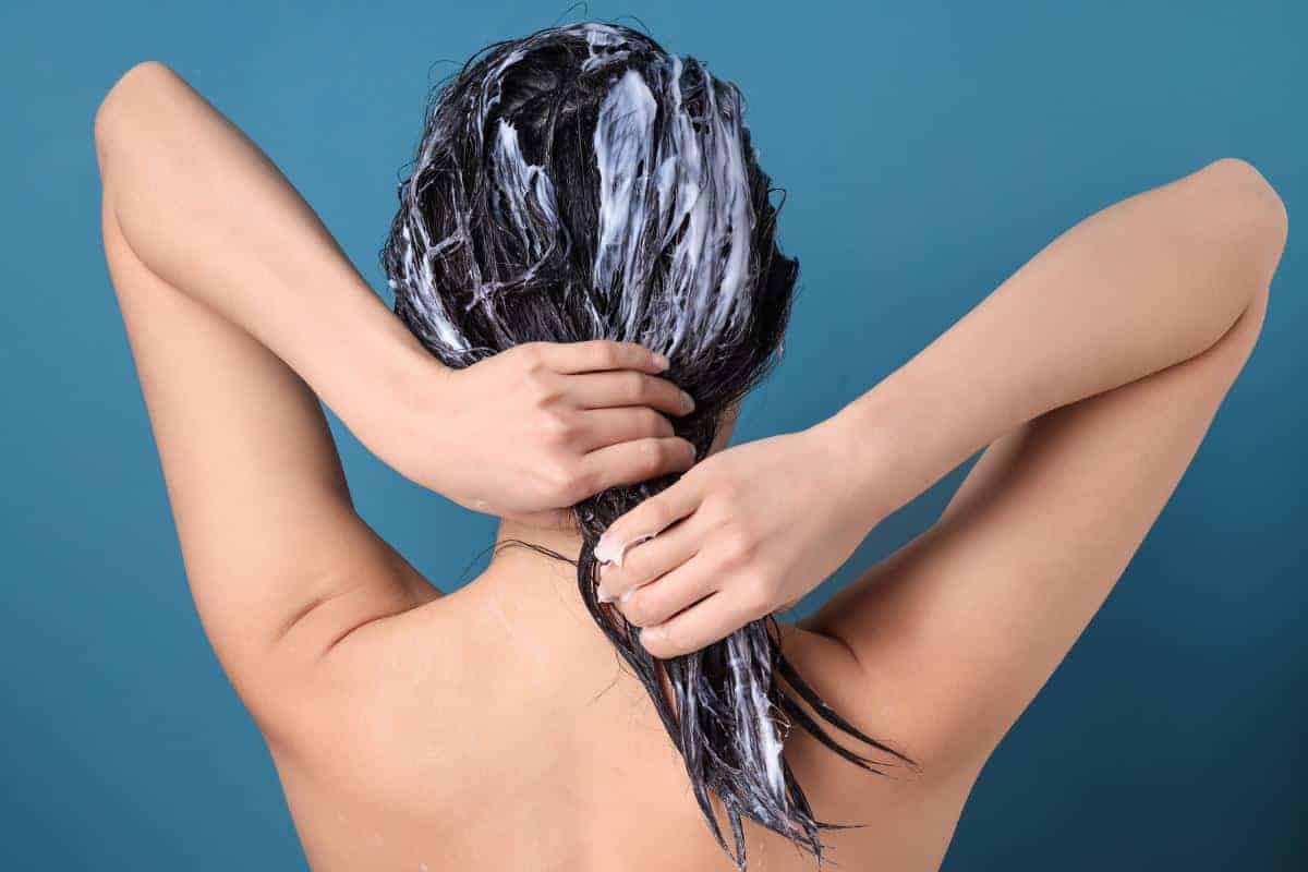 A person with wet, dark hair applies a DIY hair mask, combing it through their hair with both hands. Captured from behind against a blue background, the scene highlights self-care and pampering on a budget.