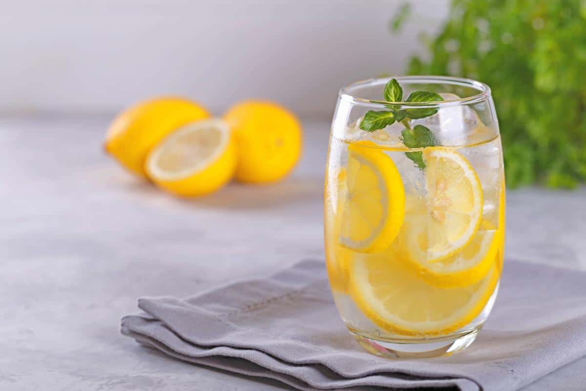 A glass of fruit-infused water with lemon slices and mint leaves sits on a grey napkin, while two whole lemons and a lemon half add a refreshing touch in the blurred background—a perfect choice among healthy drinks featuring fresh fruits.