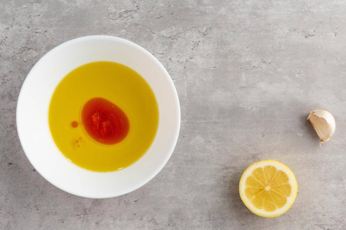 A white bowl with olive oil and a splash of red sauce sits on a gray surface next to a halved lemon and a garlic clove, perfect ingredients for a healthy orzo salad.