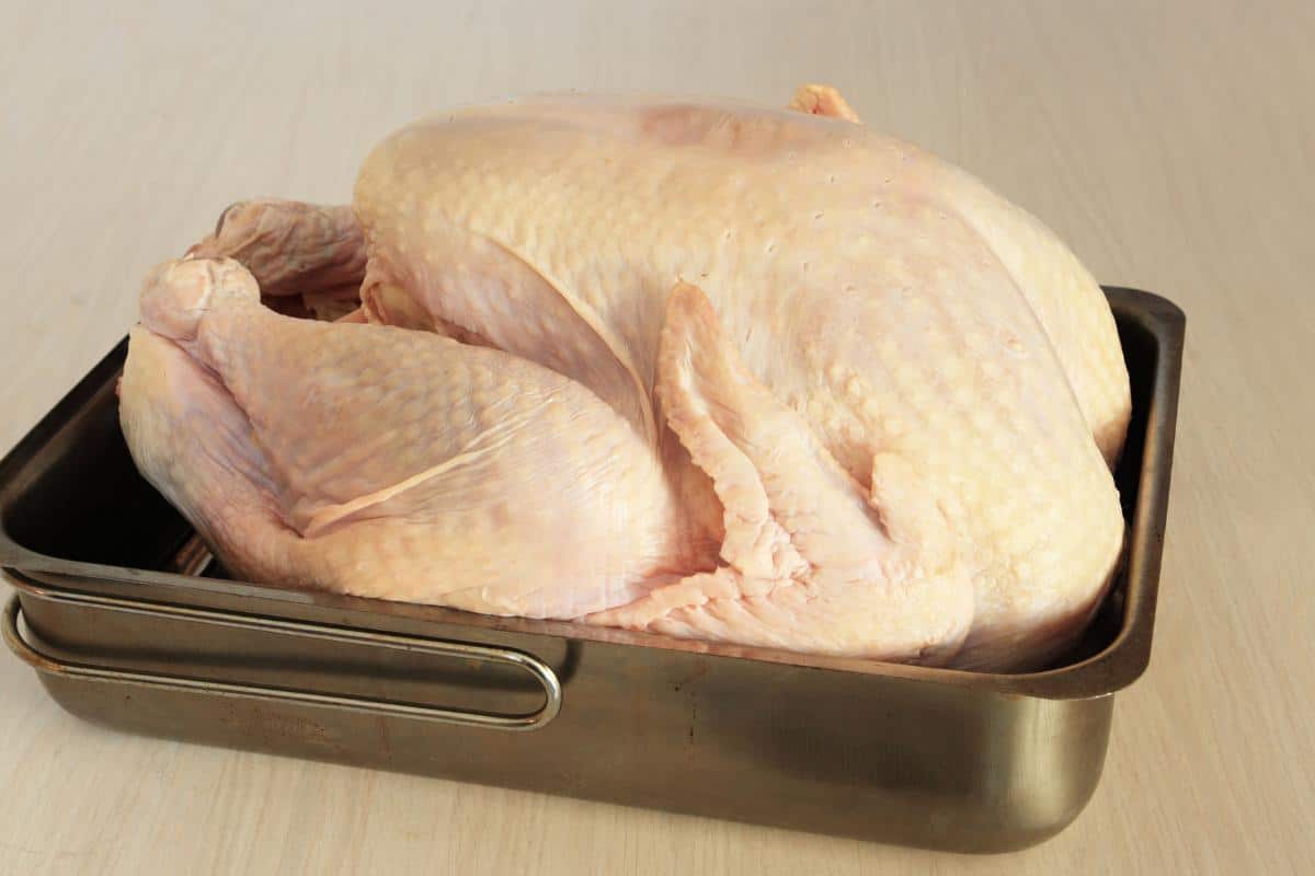 A raw whole turkey sits in a metal roasting pan on a light-colored wooden surface, ready to be prepared for cooking—perfect for those seeking a UK guide on how long to defrost turkey.