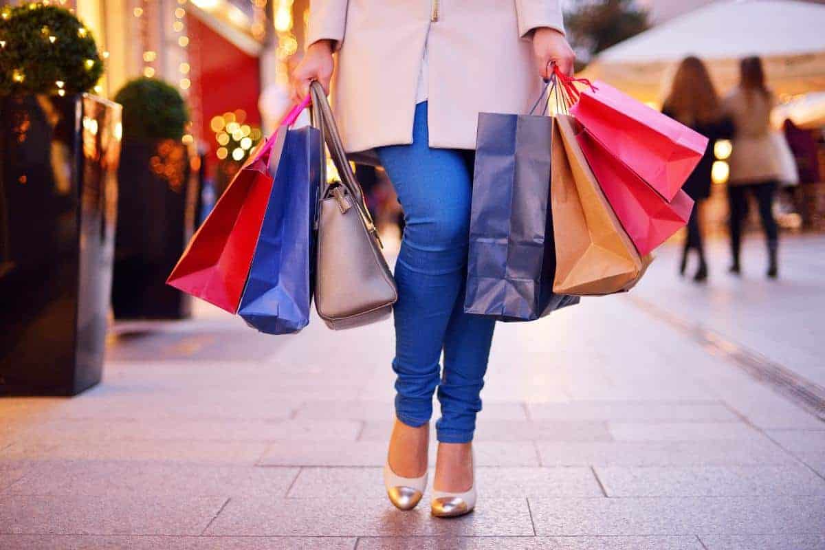A person in blue jeans and high heels is walking down a city street holding multiple colourful shopping bags, embracing the idea of shopping as exercise, with other shoppers and storefronts visible in the background.