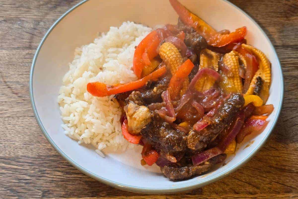 A white bowl containing steamed white rice and Ultimate Sticky Beef stir-fried with red onions, baby corn, and red bell peppers, placed on a wooden surface—a perfect Healthy Fakeaway meal.