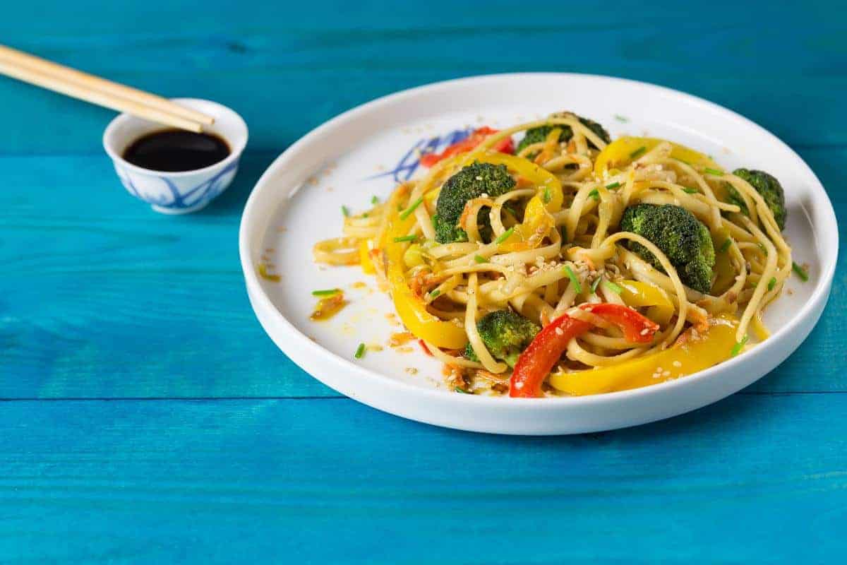 A plate of noodles with broccoli and colourful peppers, topped with sesame seeds with a pot of one of the best stir-fry sauces you could make at home.
