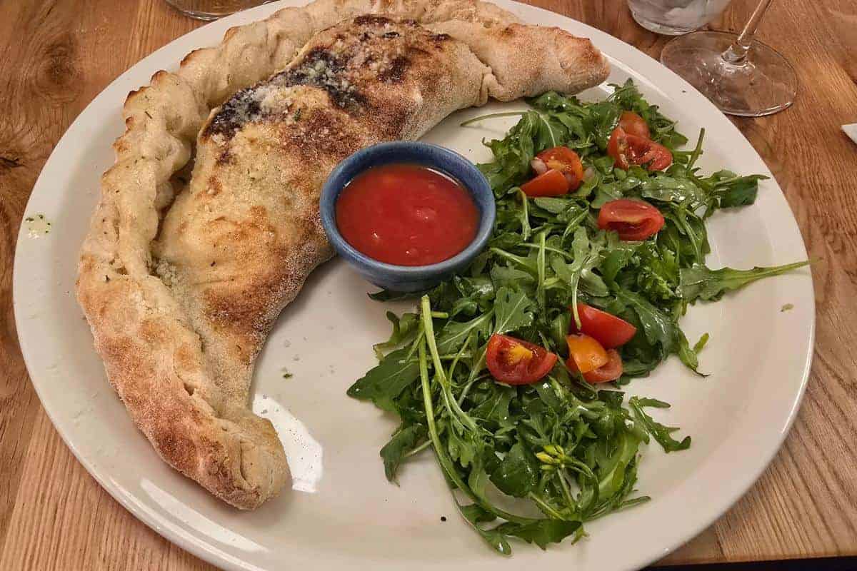 A plate with a baked calzone, a small bowl of red marinara sauce, and an arugula and cherry tomato salad, served on a wooden table at Prezzo Italian Nottingham—perfect for your next review.