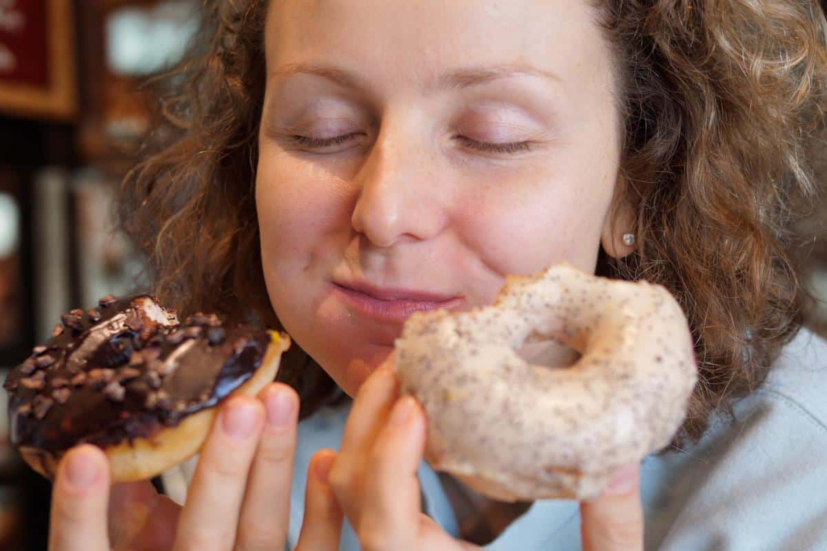 A person with curly hair and closed eyes smiles contentedly while holding a chocolate donut in one hand and a powdered donut in the other, enjoying their sweet treats—an image that may resonate with those affected by binge eating disorder.