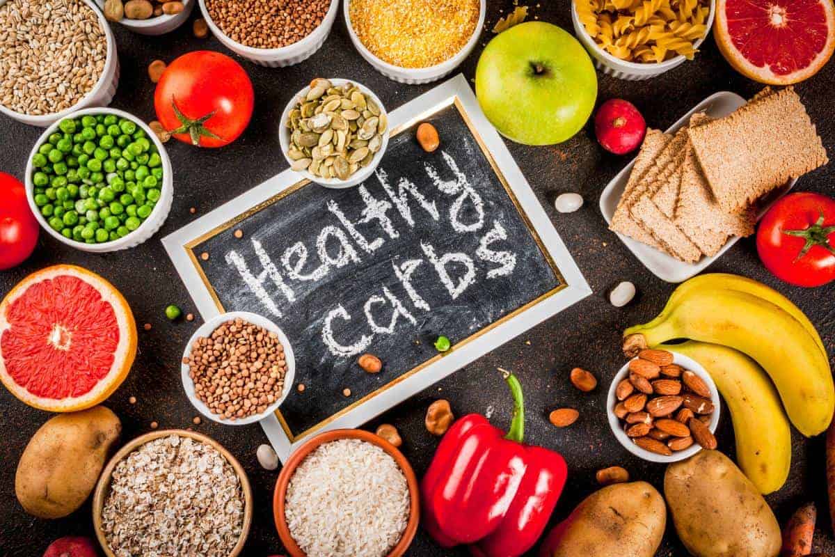 A chalkboard with “Healthy carbs” written on it is surrounded by fruits, vegetables, whole grains, seeds, nuts, potatoes, and pasta—showcasing good carbs that support balanced nutrition and can aid in weight loss.