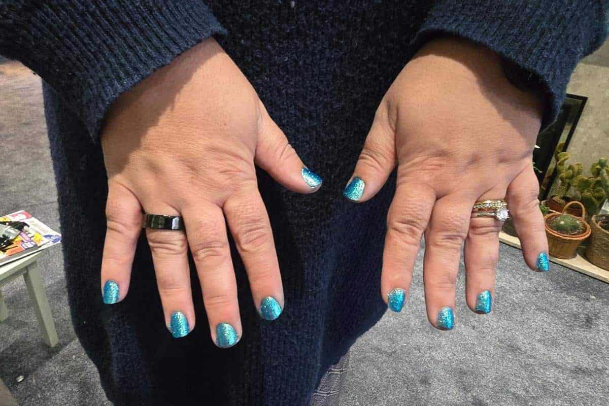 A person wearing a dark blue sweater displays their hands with short, blue glittery manicured nails and several rings, including the Ultrahuman Ring Air, on both hands. The background features a gray carpet, plants, and a small white table.