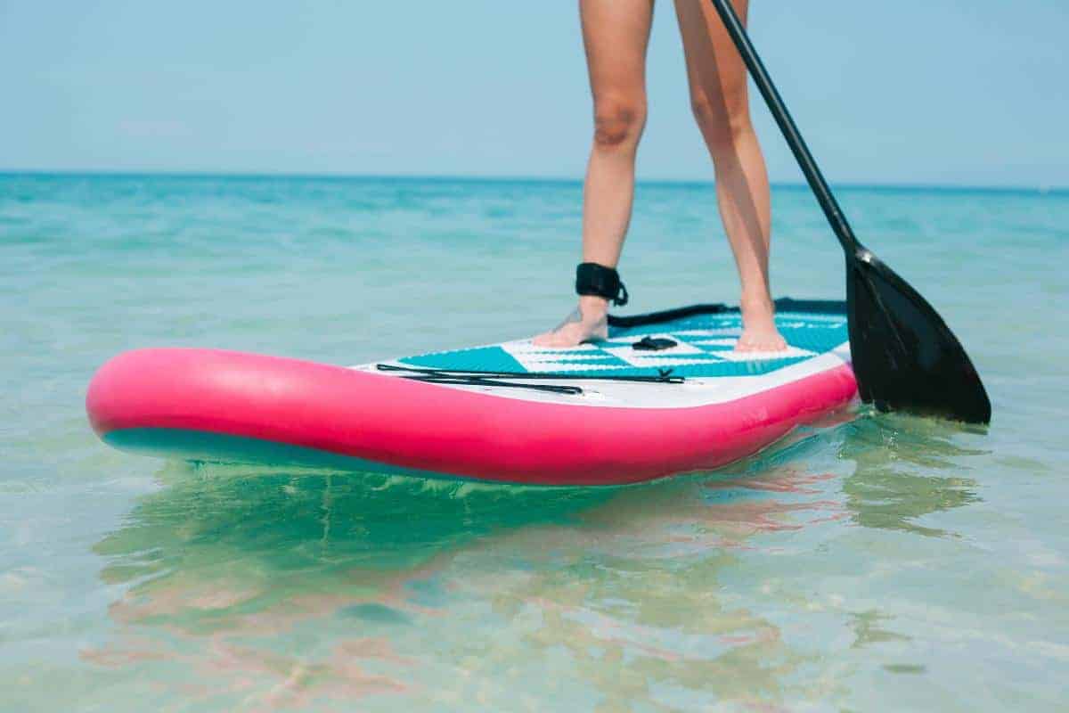 A person stands on a paddleboard with a pink edge, holding a paddle in shallow, clear blue water under a bright sky. Perfect for those seeking a beginner’s guide to paddleboarding on their next beach holiday. Only their legs and feet are visible.