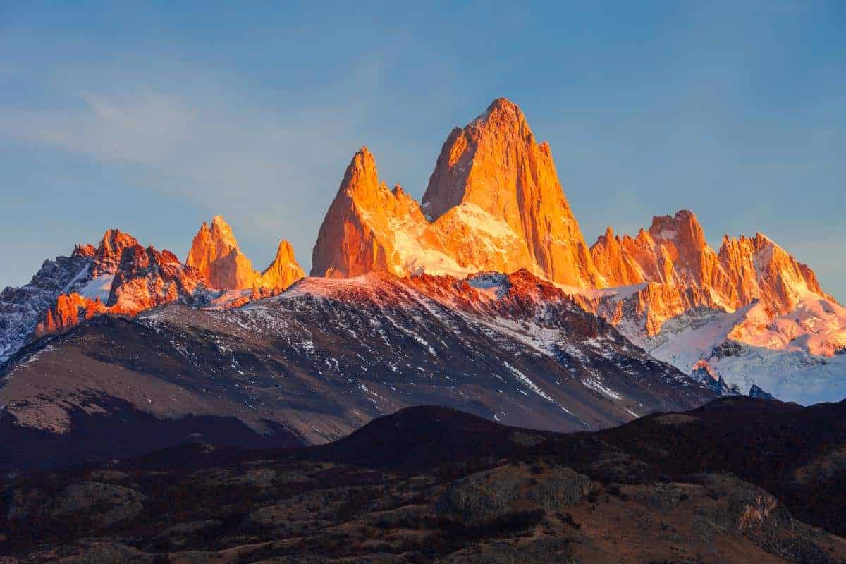 Jagged mountain peaks, topped with snow, glow orange in the warm light of sunrise or sunset. The lower slopes are shadowed and rugged, with patches of forest and rocky land in the foreground.