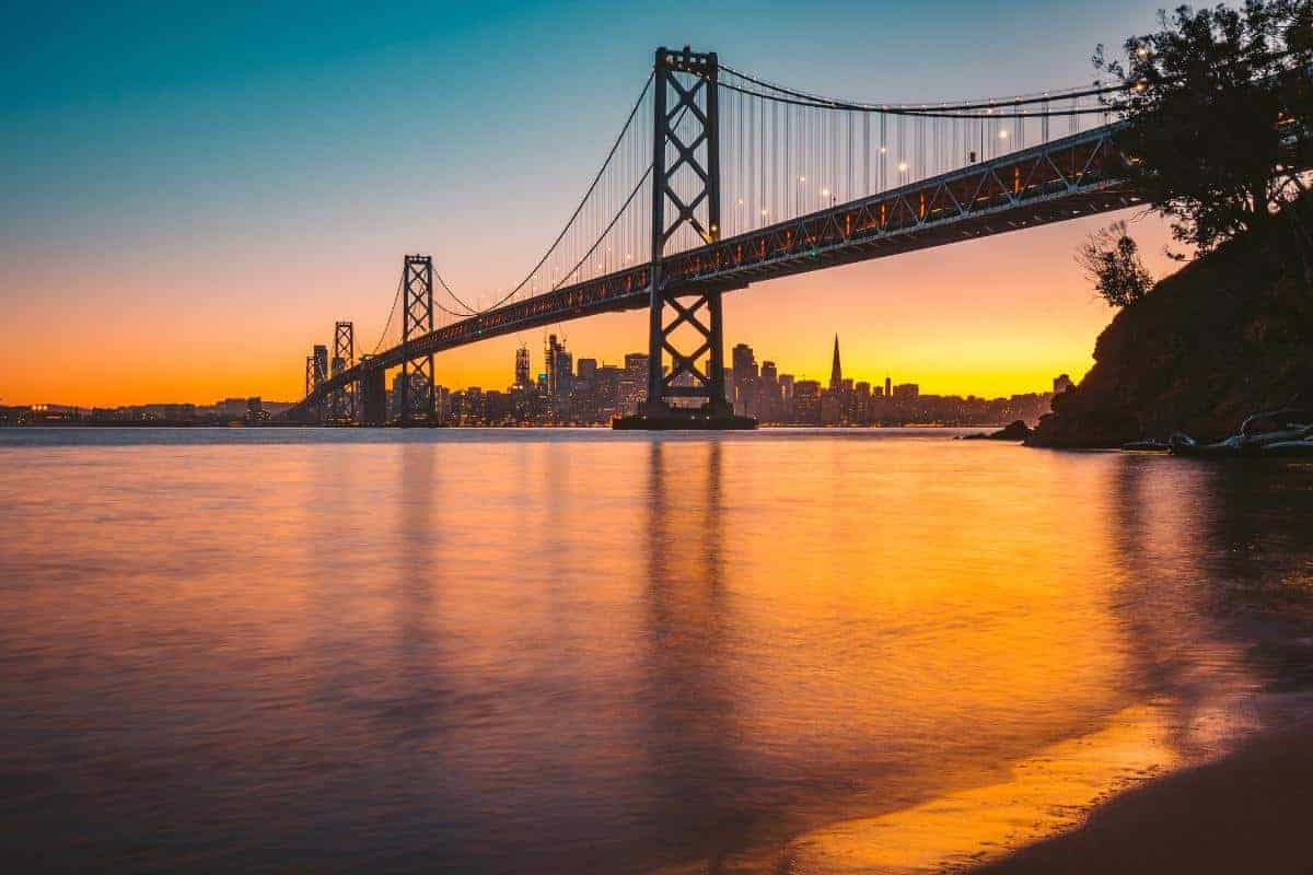 The Bay Bridge stretches over calm water at sunset, with the San Francisco skyline visible in the background and vibrant orange, yellow, and blue hues reflecting on the surface—a stunning way to get around and travel in style.