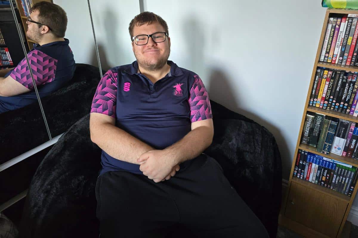 A smiling man with glasses sits on a Monster Bean Bag, wearing a navy and pink sports shirt. A mirrored wardrobe and a bookshelf filled with DVDs and books are visible in the background.