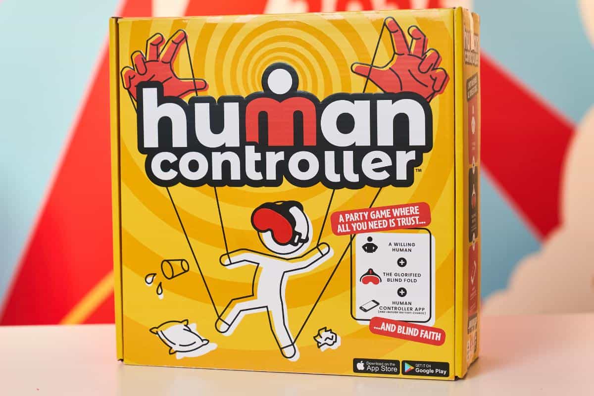 A yellow game box labeled "Human Controller" features a cartoon figure being controlled by puppet strings and large red hands above. Perfect for kids, this party game is sure to be on every Christmas List of Top Toys 2025, with fun depictions of game elements.