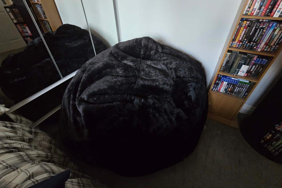 A large black Monster Bean Bag from GreatBeanBags sits in the corner of a room next to mirrored wardrobe doors, a bed with plaid bedding, and bookshelves filled with DVDs and books.