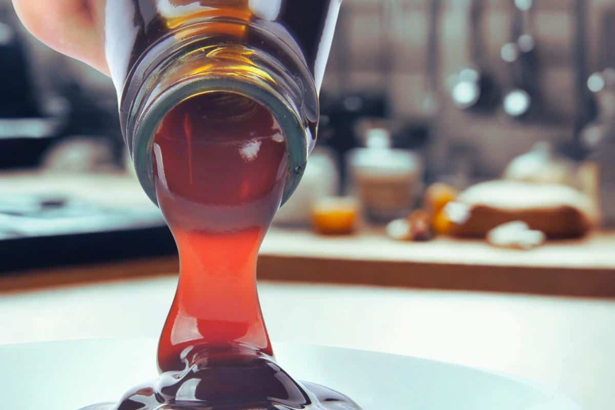 A close-up of vegan syrup being poured from a glass bottle onto a white plate, with a blurred kitchen background—perfect as one of the best vegetarian oyster sauce alternatives.