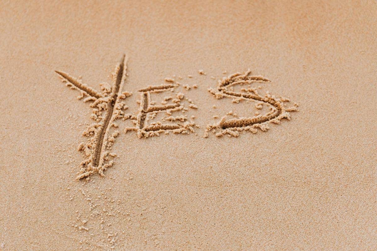The word "YES" is written in capital letters in the sand, with smooth and rough edges, on a light brown sandy surface, capturing the Power of Saying Yes and its potential to Change Your Life.