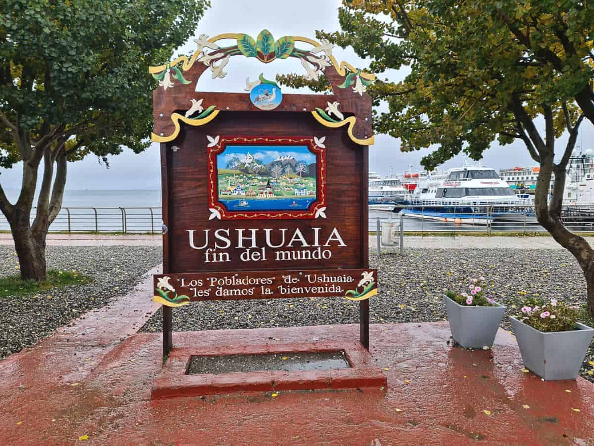 Дървена табела с текст Ushuaia Fin del mundo (Ушуая - край на света на английски език) между две дървета на пътническия кей в Ушуая и няколко катамарана на заден план вдясно