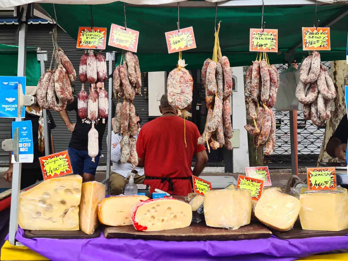 At a bustling food stall at Mataderos Fair, various cured meats hang above while an assortment of large cheese wheels is prominently displayed on the table in front. Brightly colored signs advertise prices and specials for items such as "picada," "salamin," and "queso." The scene is indicative of a local market, with vendors and shoppers engaged in the vibrant commerce of traditional food products.