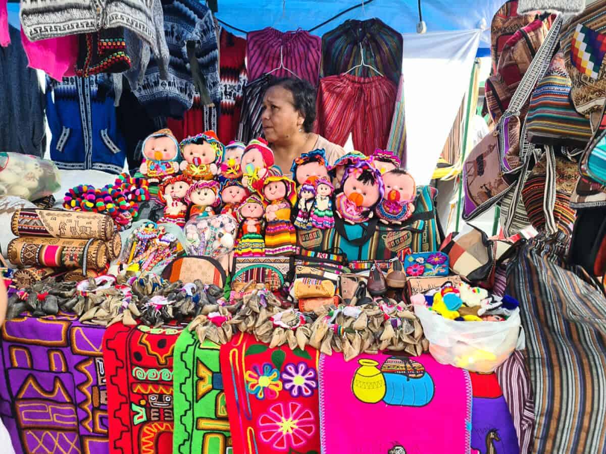 A woman selling colorful handcrafted items at Mataderos market. The merchandise includes traditional dolls dressed in vibrant, multicolored outfits, woven blankets with intricate patterns, and various bags with ethnic designs. The items seem to reflect a rich cultural heritage, likely showcasing local craftsmanship and textiles. The scene is bustling with a diverse array of goods, indicating a lively local market atmosphere.