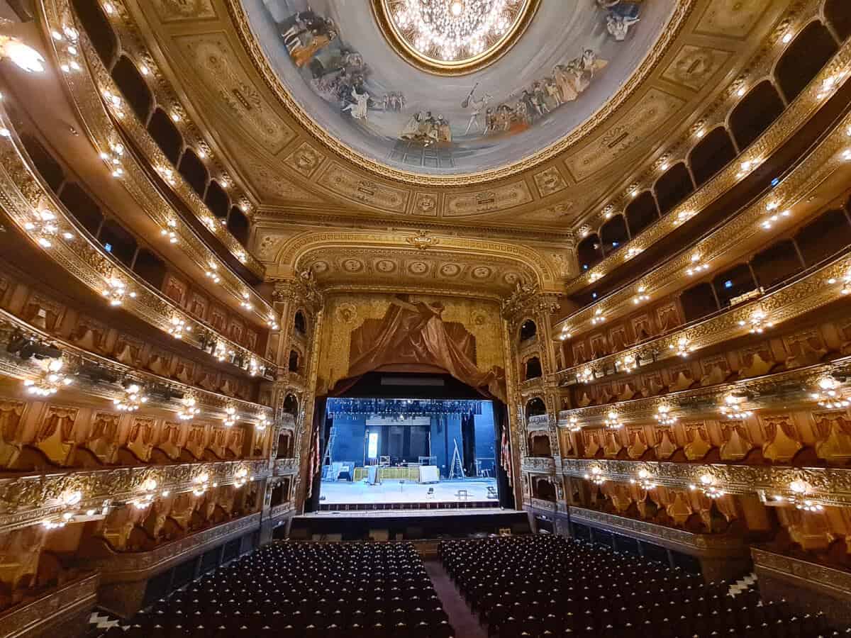 This image captures the interior of Teatro Colón's auditorium in Buenos Aires, renowned for its opulent design and exceptional acoustics. The photograph shows the ornate golden balconies stacked in a horseshoe arrangement, classic to traditional European opera houses. Each balcony level is adorned with intricate golden embellishments and plush red velvet seating. The ceiling features a magnificent fresco, surrounded by elaborate moldings, adding to the grandeur of the space. The stage is visible in the background with its grand curtain partially drawn, suggesting preparations for a performance. The overall ambience is of historical elegance and theatrical splendor.