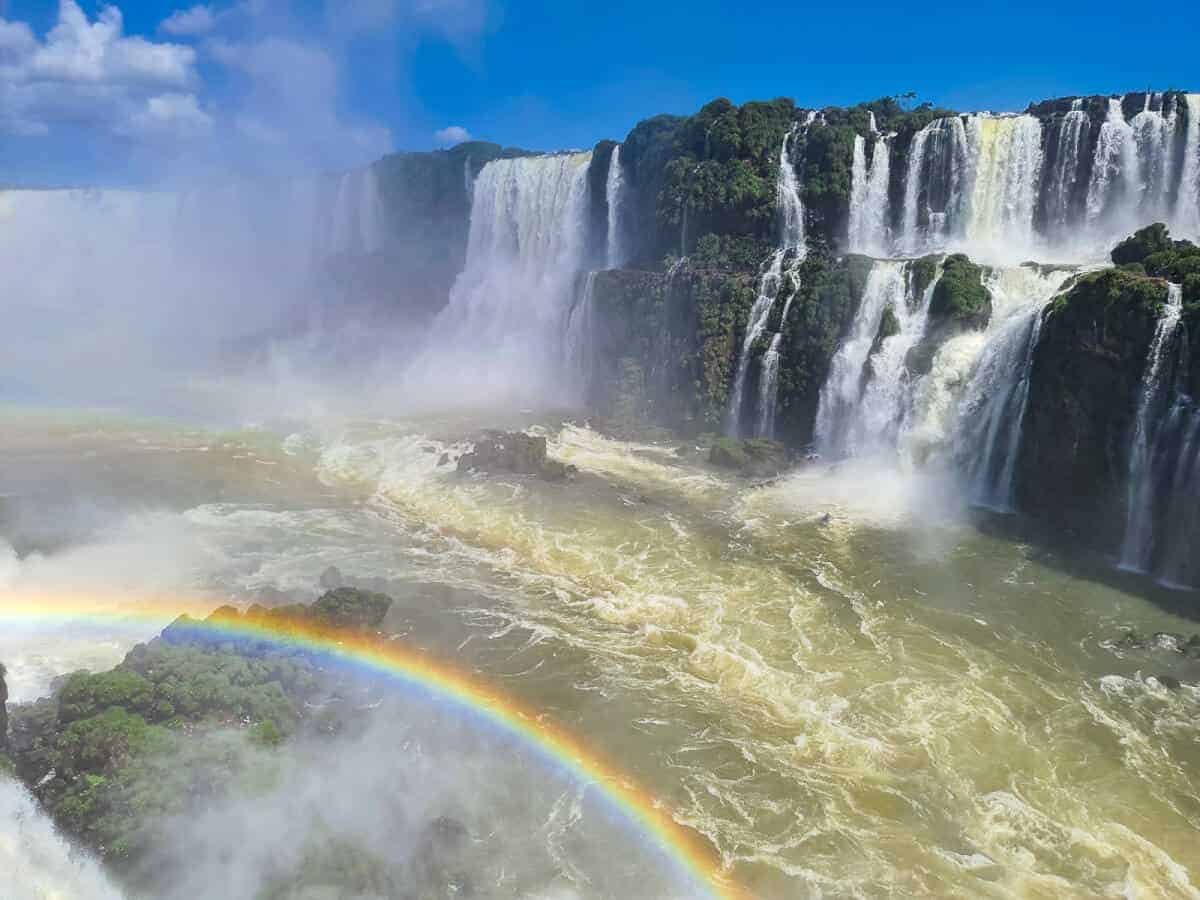 The photograph showcases a striking view of Iguazú Falls, where numerous waterfalls cascade into a turbulent, golden-brown river. A vivid rainbow arches gracefully through the mist created by the falls, adding a spectrum of color to the scene. The lush greenery of the surrounding forest is partially visible, framing the natural wonder, while the powerful rush of water conveys a sense of the falls' majesty and the dynamic energy of nature.