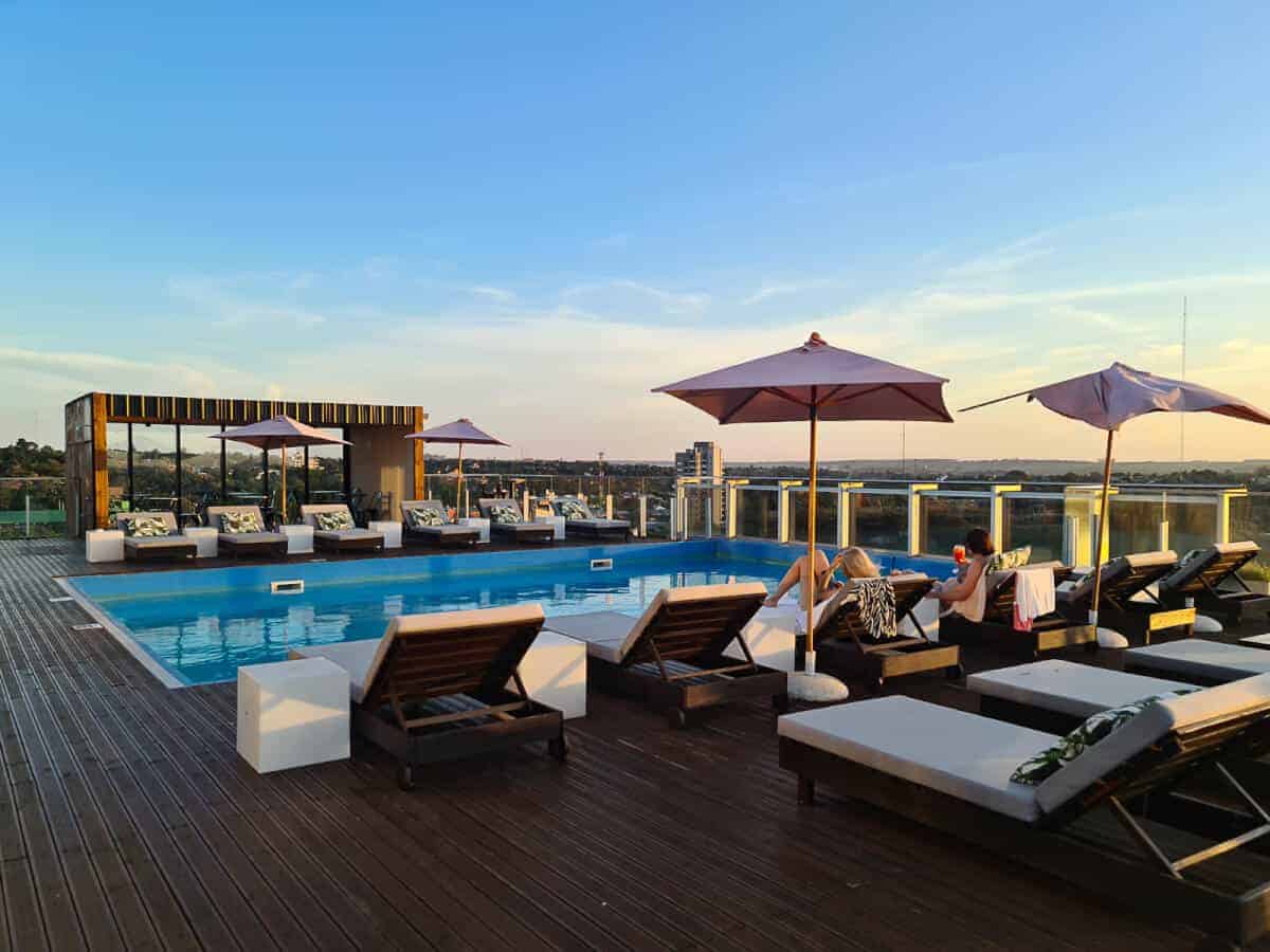 The image shows a luxurious rooftop swimming pool at dusk, with a clear blue sky above. The pool area is adorned with stylish sun loungers, some of which are occupied by guests enjoying the tranquil setting. Large umbrellas provide shade, and a chic bar structure offers refreshments. The wooden deck and glass balustrades add to the modern aesthetic, while the background reveals a sprawling landscape that fades into the horizon, highlighting the urban setting of the O2 Iguazu Hotel.