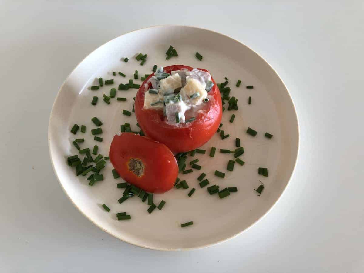 Cold stuffed tomato in a white plate surrounded by chopped chives.