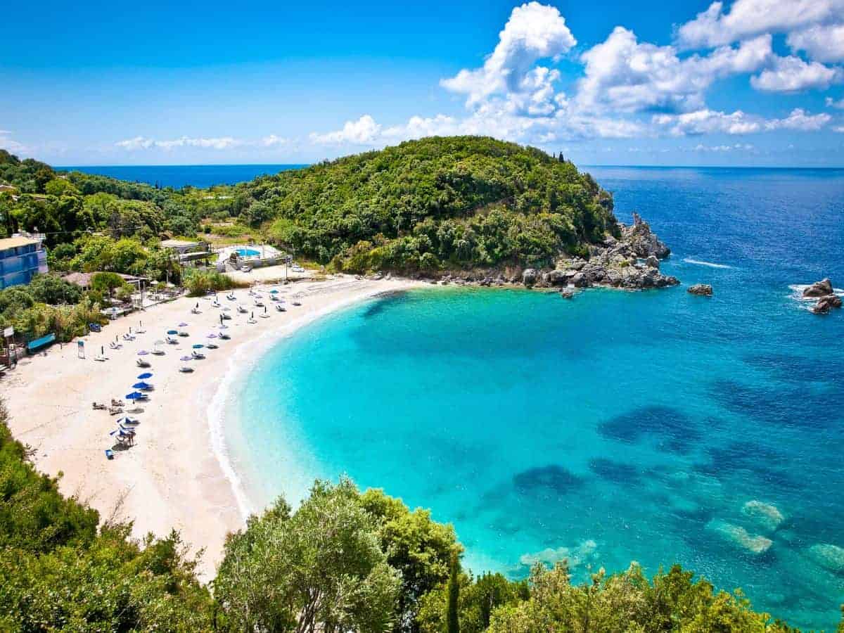 Best Beaches in Milos, Greece. White Sand Beach Best beaches in Milos. Image of white sandy curved beach with green tree covered spit at the far right, surrounded to the right with clear turqoise sea. Blue sky and white clouds above and a few sunbeds and blue parasols on the sand.