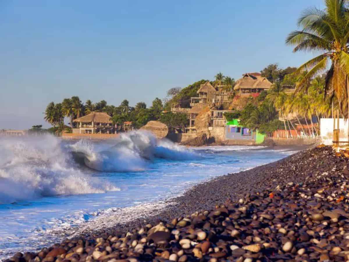 Things to do in Playa El Tunco. Things to do in Playa El Tunco. Image of pepple beach with big surf waves crashing on the shore. To the right on the land are palm trees and palm leaf roof buildings.