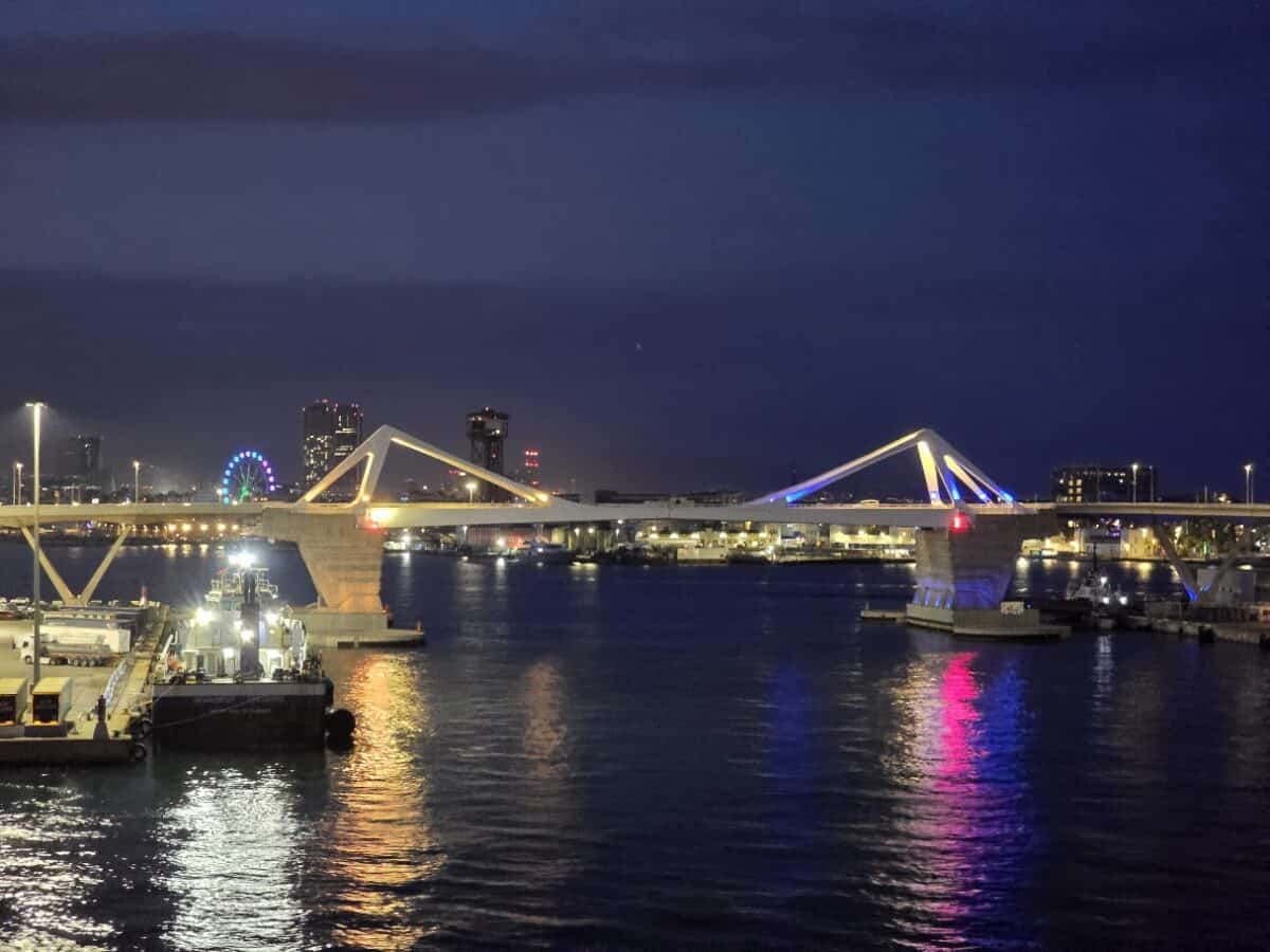 A modern bridge illuminated with colorful lights spans across a river at night, reflecting on the water; city buildings and a lit Ferris wheel evoke the vibrant atmosphere of an overnight journey from Barcelona to Palma.