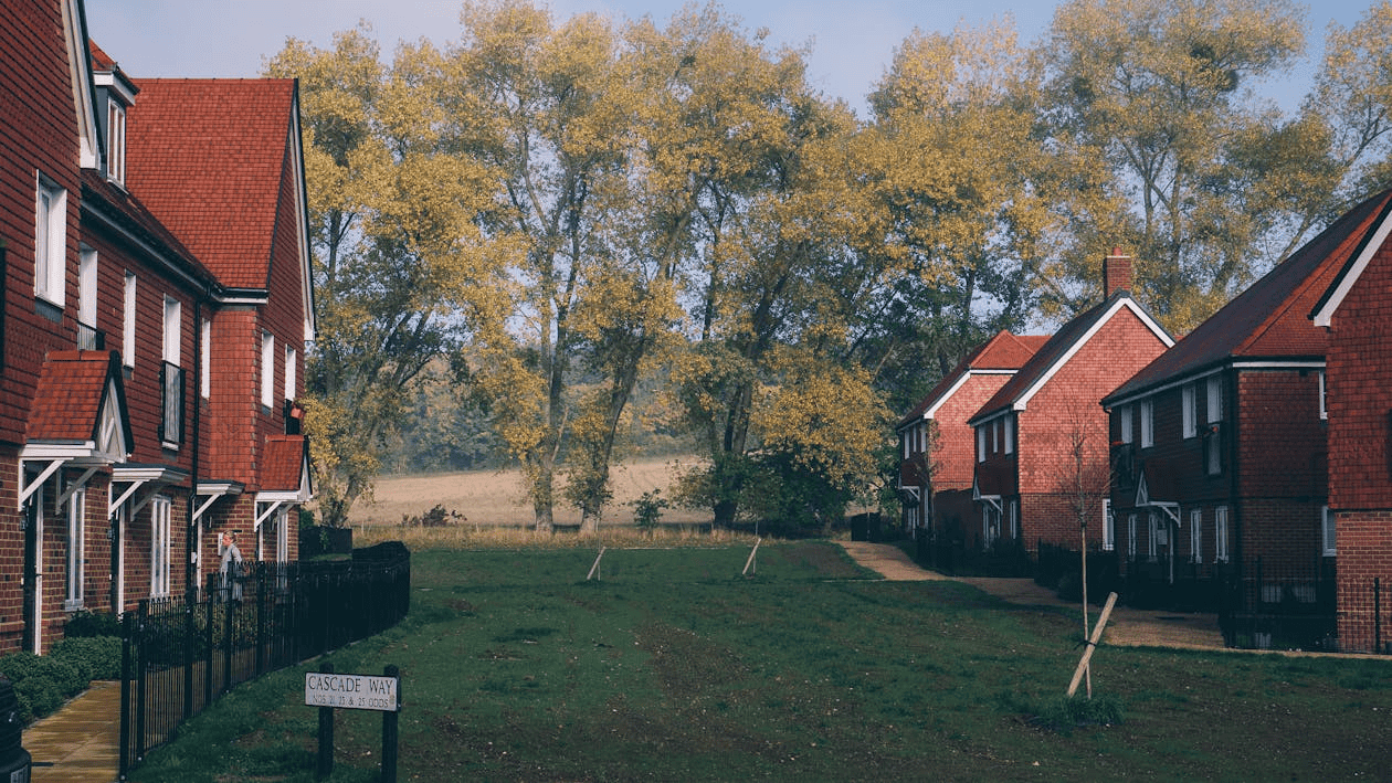 A quiet residential street lined with red-brick houses, bordered by a grassy area and tall trees with autumn foliage under a clear sky. A sign reading "Cascade Way" is visible in the foreground.