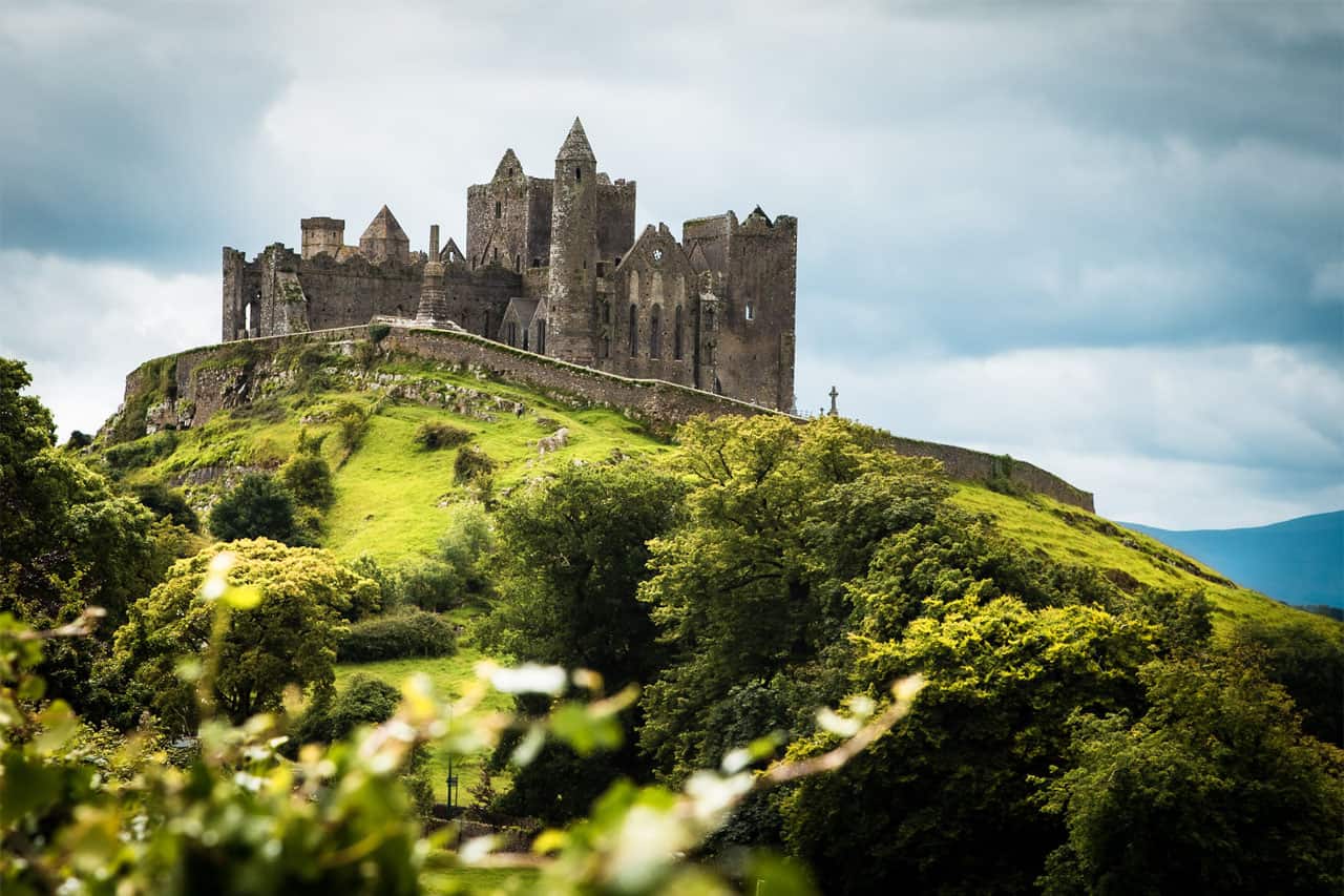 The Rock of Cashel, a historic medieval fortress, stands majestically atop a lush green hill in County Tipperary, Ireland. The stone structure features ancient towers and crumbling walls, surrounded by a stone barrier, with dense trees and rolling hills in the foreground. A cloudy sky looms above, adding a dramatic backdrop to the iconic landmark.