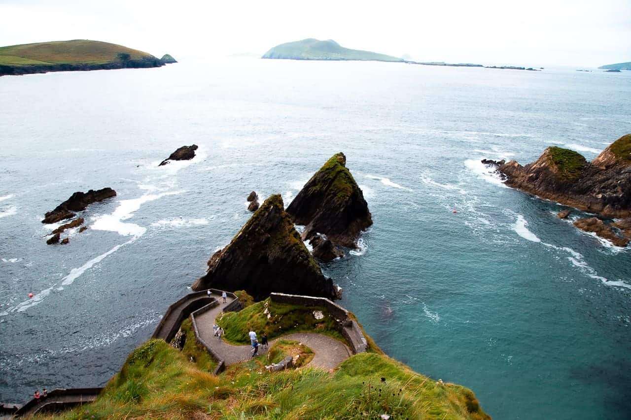 A scenic view of Dunquin Pier on the Dingle Peninsula in Ireland, featuring a winding stone pathway leading down to sharp, jagged sea stacks rising from the Atlantic Ocean. The turquoise water gently laps against the rocky shoreline, and distant green hills can be seen on the horizon under a cloudy sky. A few people are walking along the path, enjoying the breathtaking coastal landscape.
