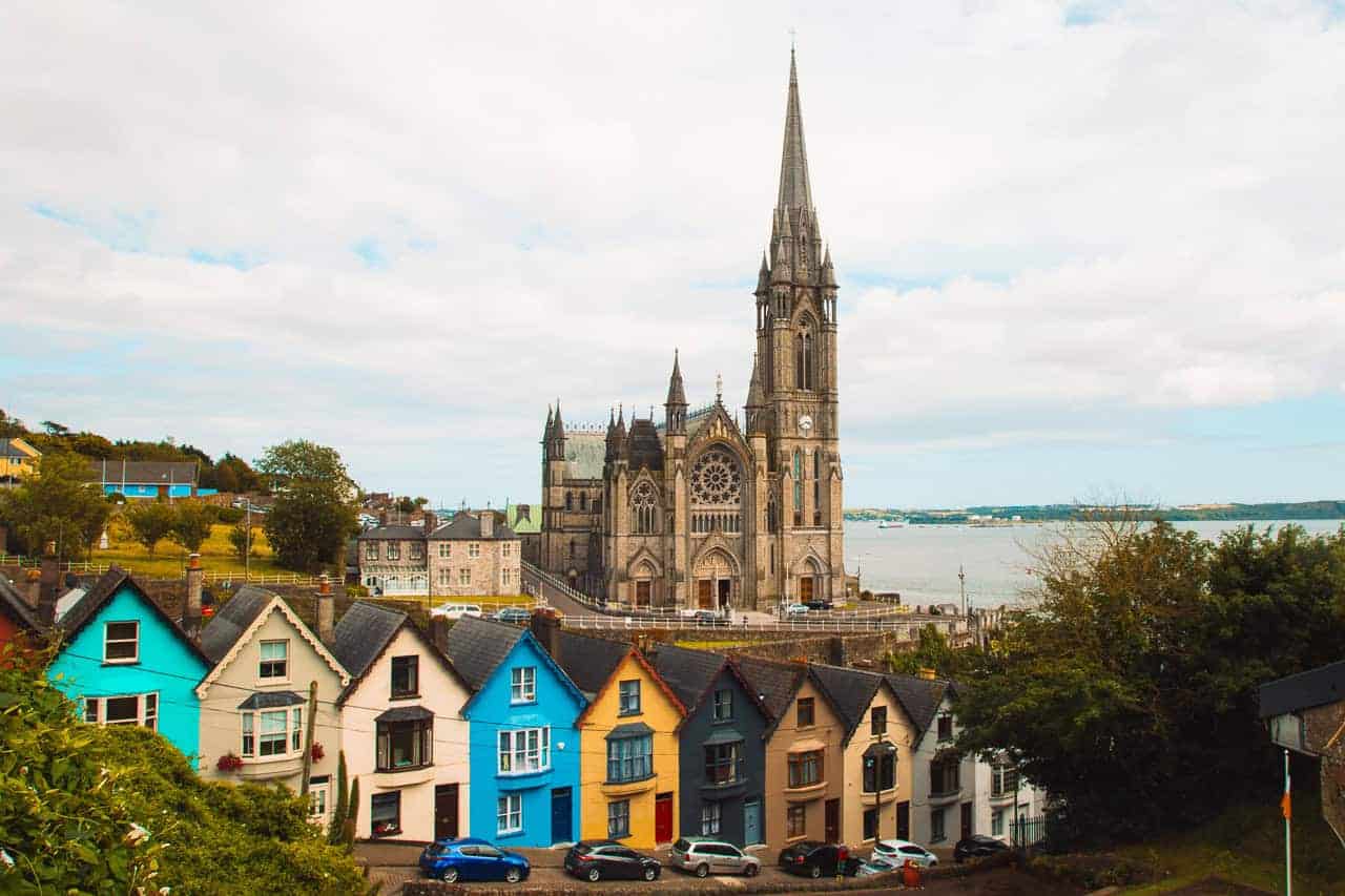 A vibrant view of colorful row houses in front of St. Colman’s Cathedral in Cobh, Ireland, with its towering spire and intricate Gothic architecture. The scene overlooks the harbor in the background under a partly cloudy sky, blending historic charm with coastal beauty.